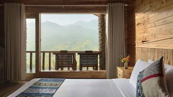 A bedroom with a large bed, wooden walls, a balcony with two chairs, and a view of mountains.