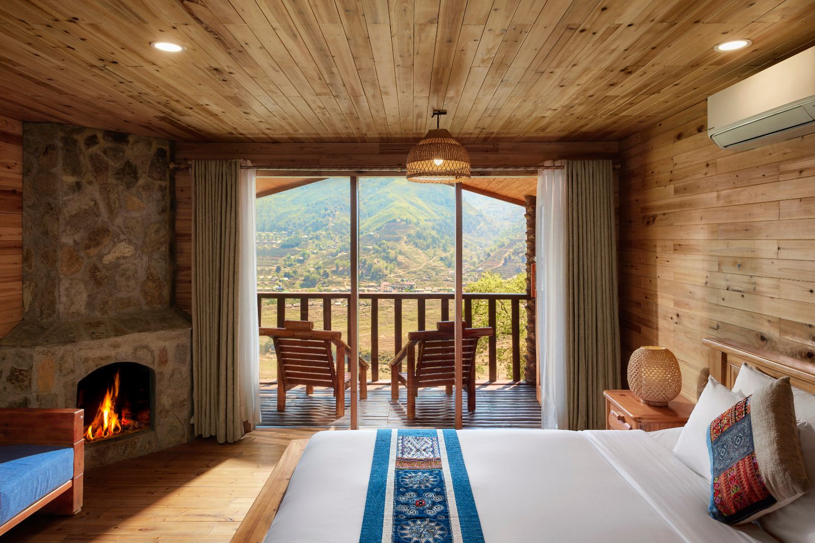 A bedroom with a stone fireplace, a large bed, a balcony with chairs, and a view of the countryside.
