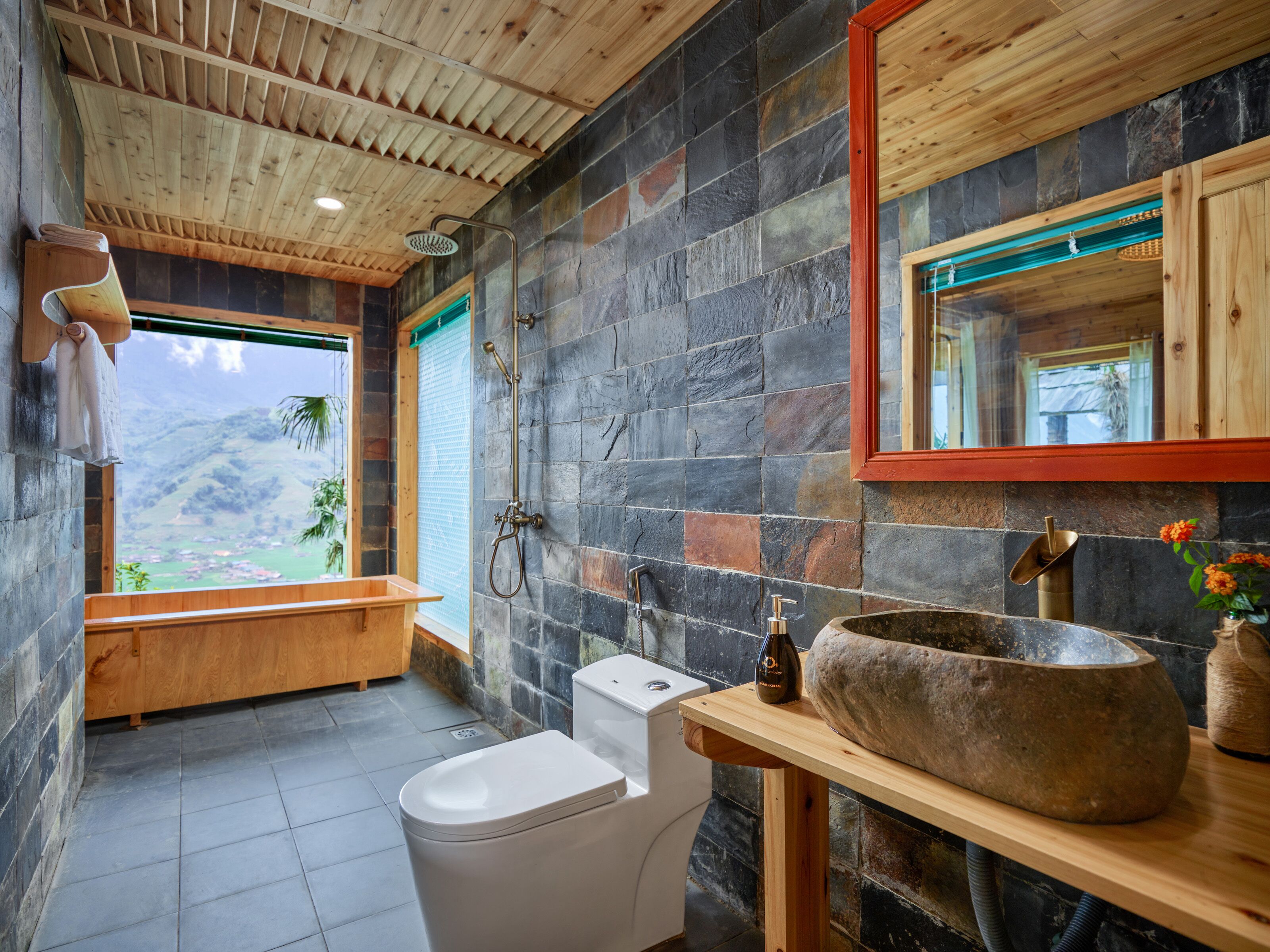 A bathroom with a stone wall, wooden ceiling, and a large window with a view of greenery.