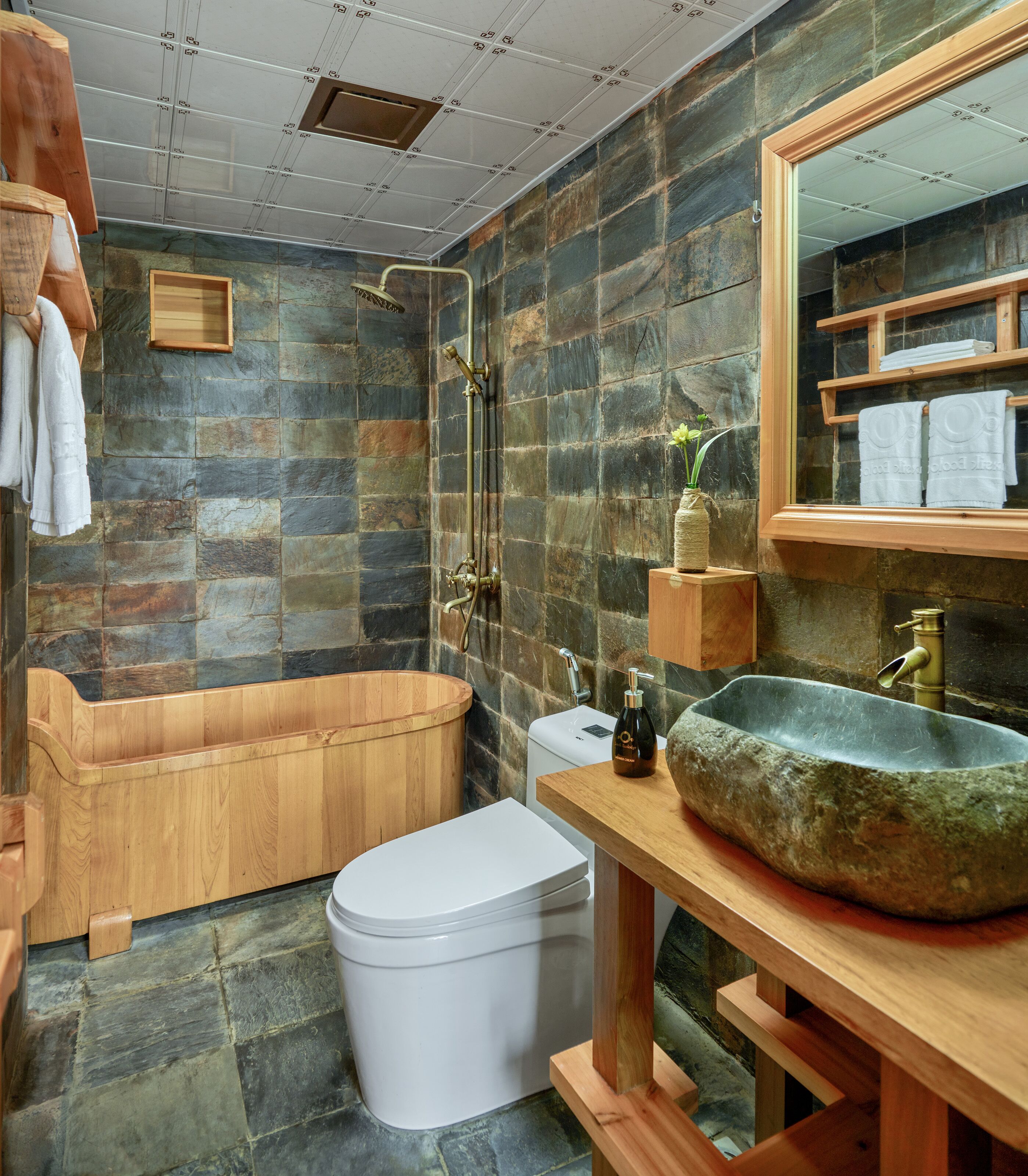 Une salle de bain avec un mur en pierre, une baignoire en bois, des toilettes blanches et un grand lavabo en pierre.