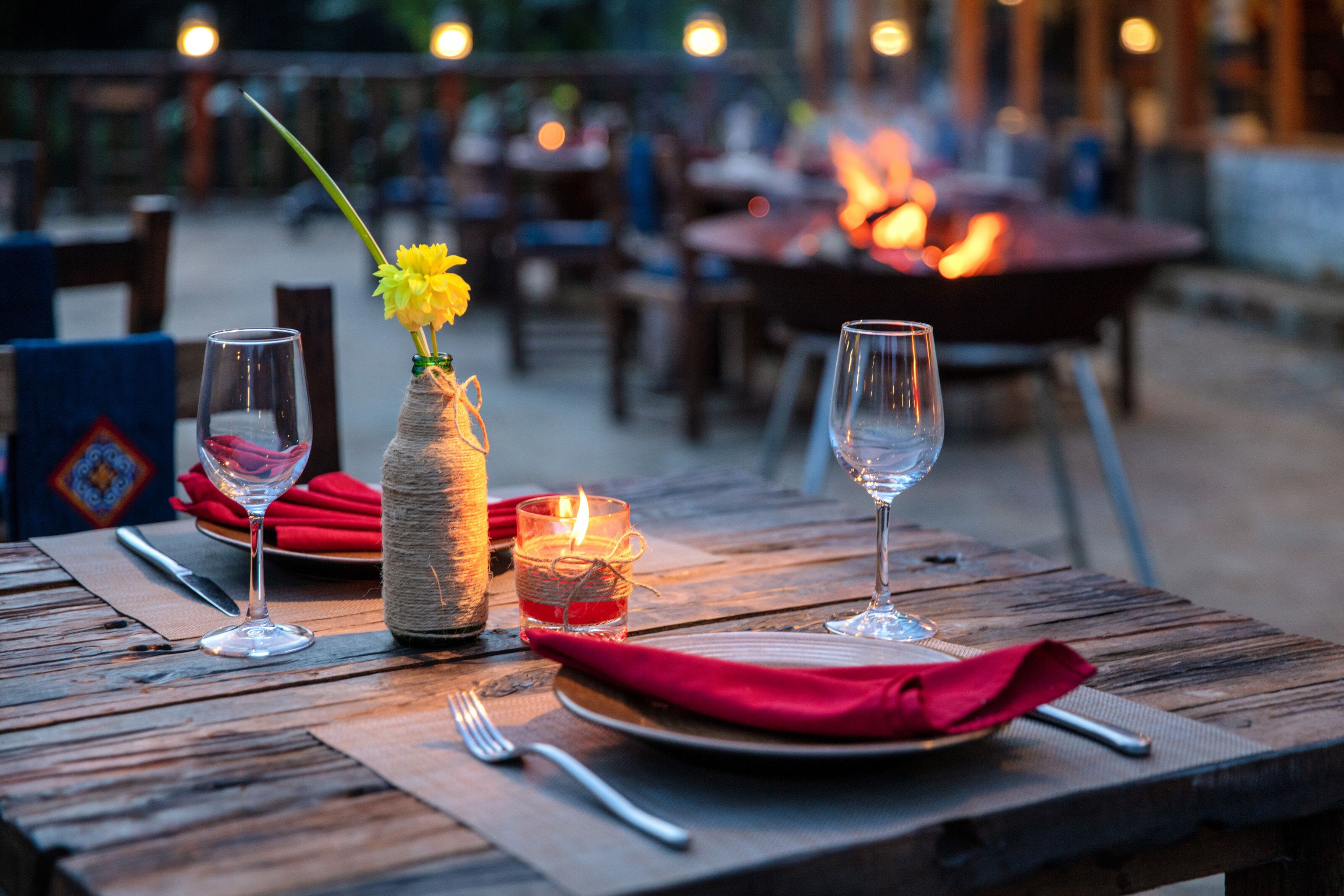 Een eettafel gedekt met wijnglazen, een vaas met een bloem en een kaars, met een vage achtergrond die een buitenomgeving suggereert.
