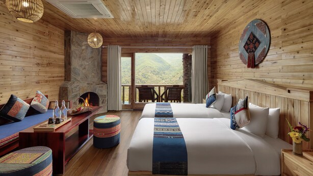 A cozy room with wooden walls and a stone fireplace. There are two beds with blue and white bedding, a wooden bench, and a table with a vase of flowers. - Laxsik Ecolodge (Sa Pa)