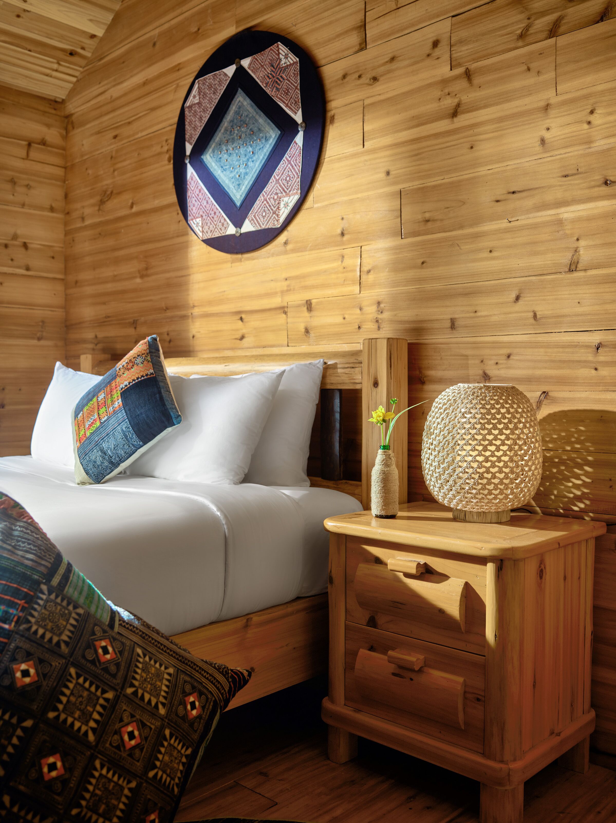 A wooden cabin interior with a bed, a nightstand, a decorative wall hanging, and a patterned throw blanket.