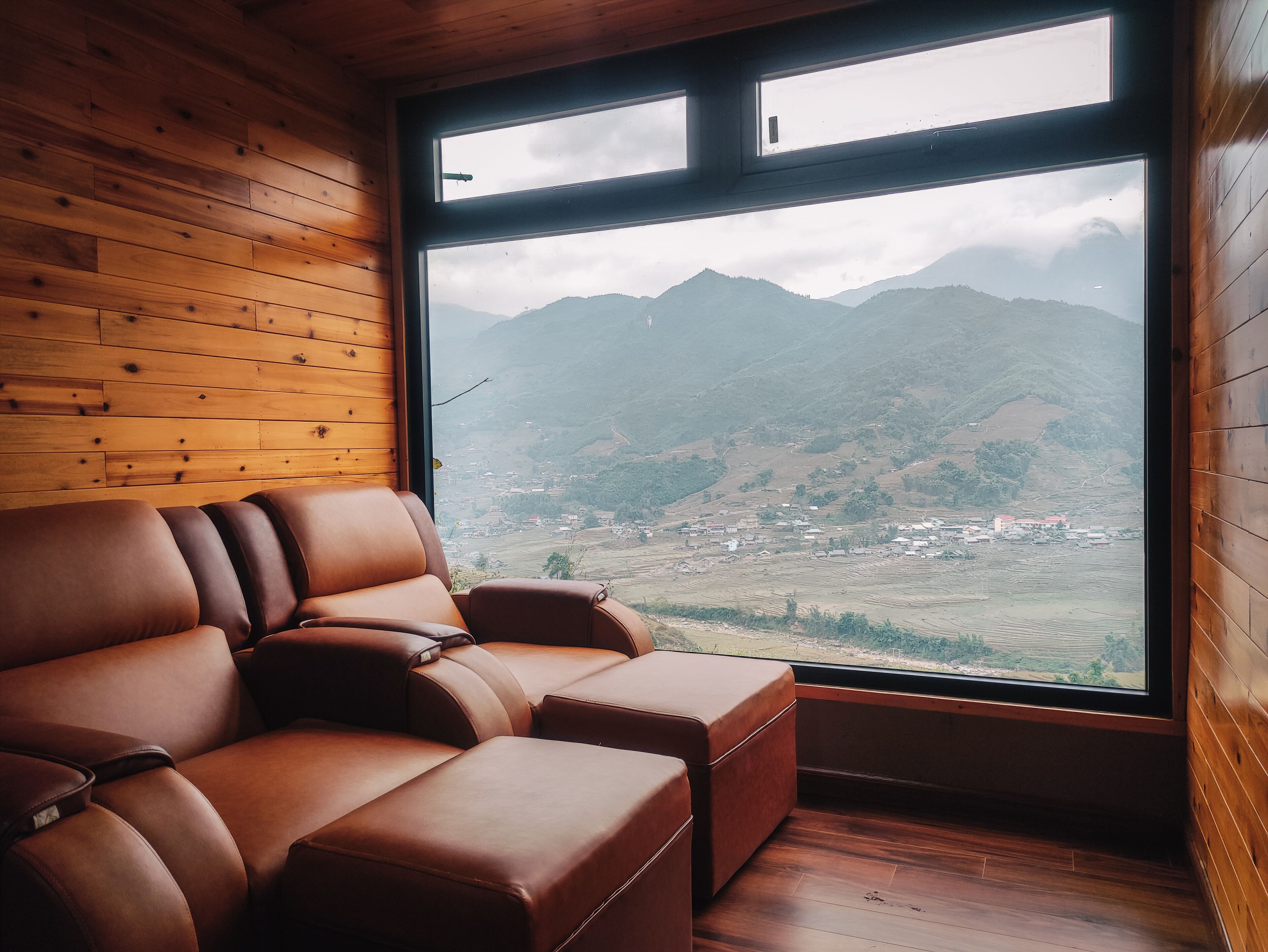 A wooden interior with a leather sofa and ottoman, a large window with a view of mountains and a village.