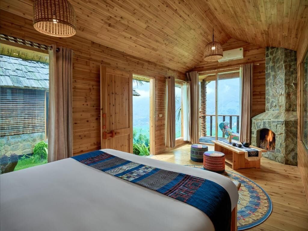 A wooden cabin bedroom with a large bed, a stone fireplace, and a view of the outdoors.