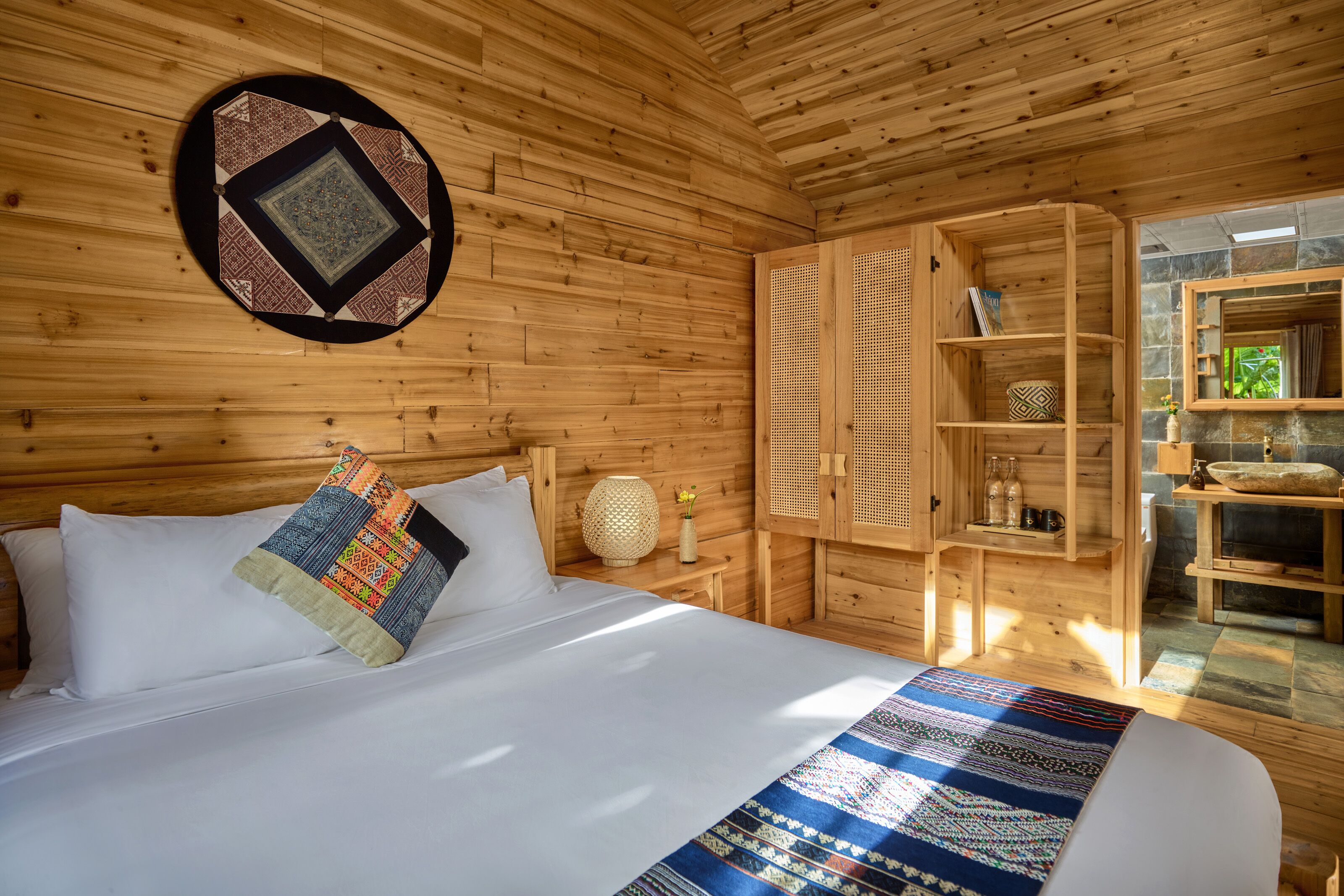 Een houten kamer met een bed, een geweven vloerkleed, een rieten mand en een plank met potten.
