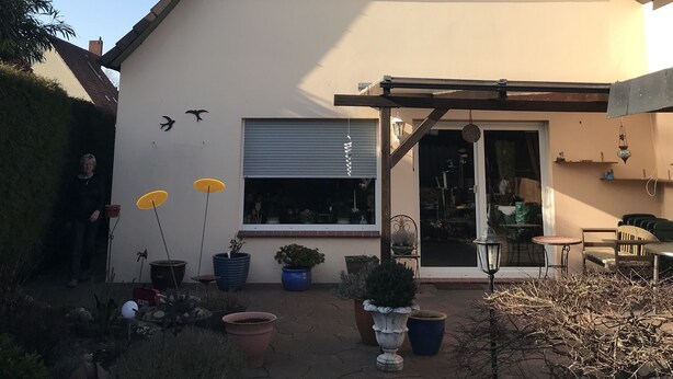 Una casa de dos pisos con techo inclinado, puerta de garaje y un patio cubierto con plantas en macetas.