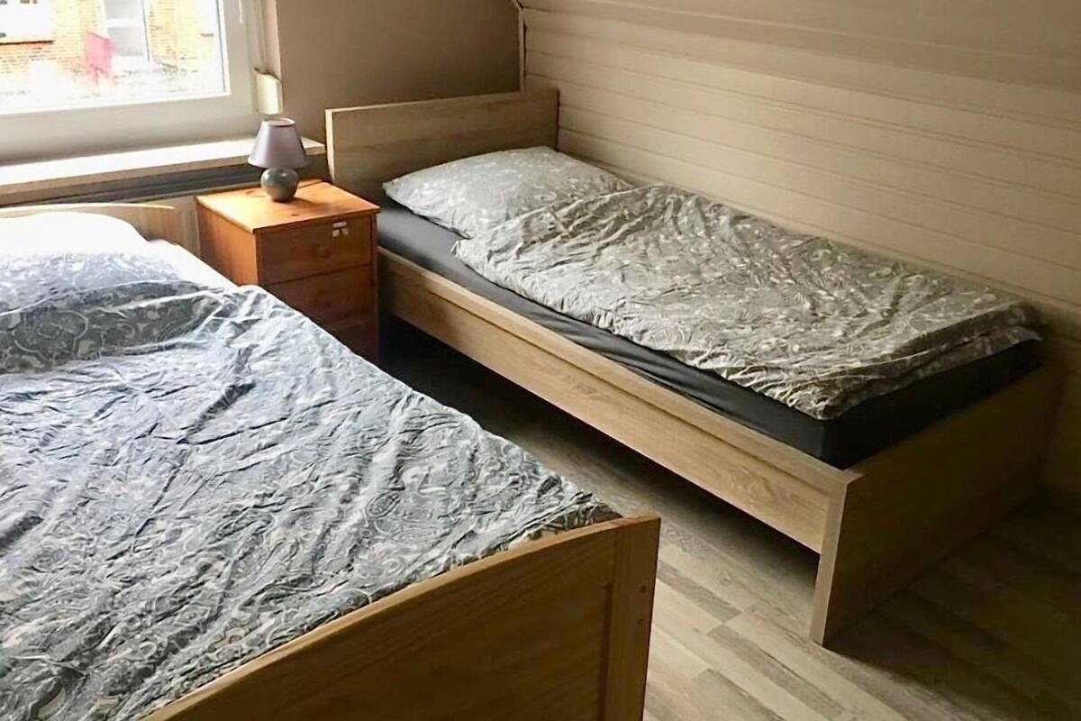 Un petit logement composé d’une seule pièce, doté d’un plafond en pente, d’un lit en bois, d’une table de chevet avec une lampe et d’une fenêtre donnant sur l’extérieur.