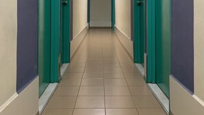 Un long couloir étroit, avec des portes de couleur turquoise de part et d’autre et un sol carrelé de couleur beige.