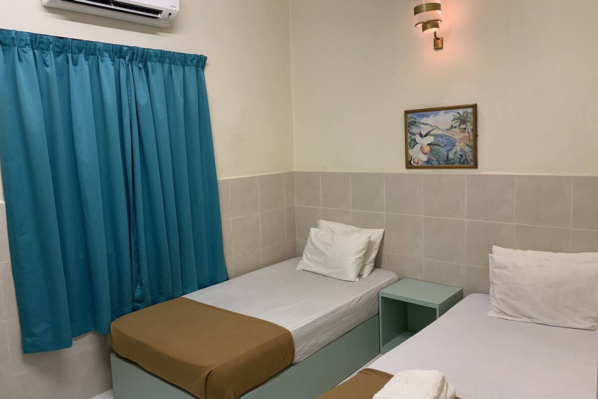 A hotel room with two beds, a blue curtain, an air conditioner, a small painting, and a bedside lamp.