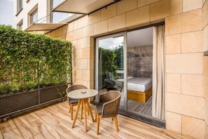 Un balcón moderno al aire libre con piso de madera, una mesita redonda pequeña, dos sillas y un ventanal de vidrio que da a un dormitorio con cama.