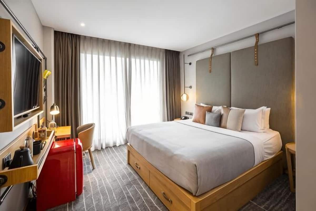 A hotel room with a bed, a desk, a chair, a TV, and a large window with curtains.