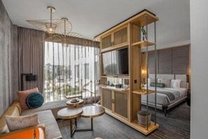 A modern hotel room with a sofa, a dining table, a TV, and a large window.