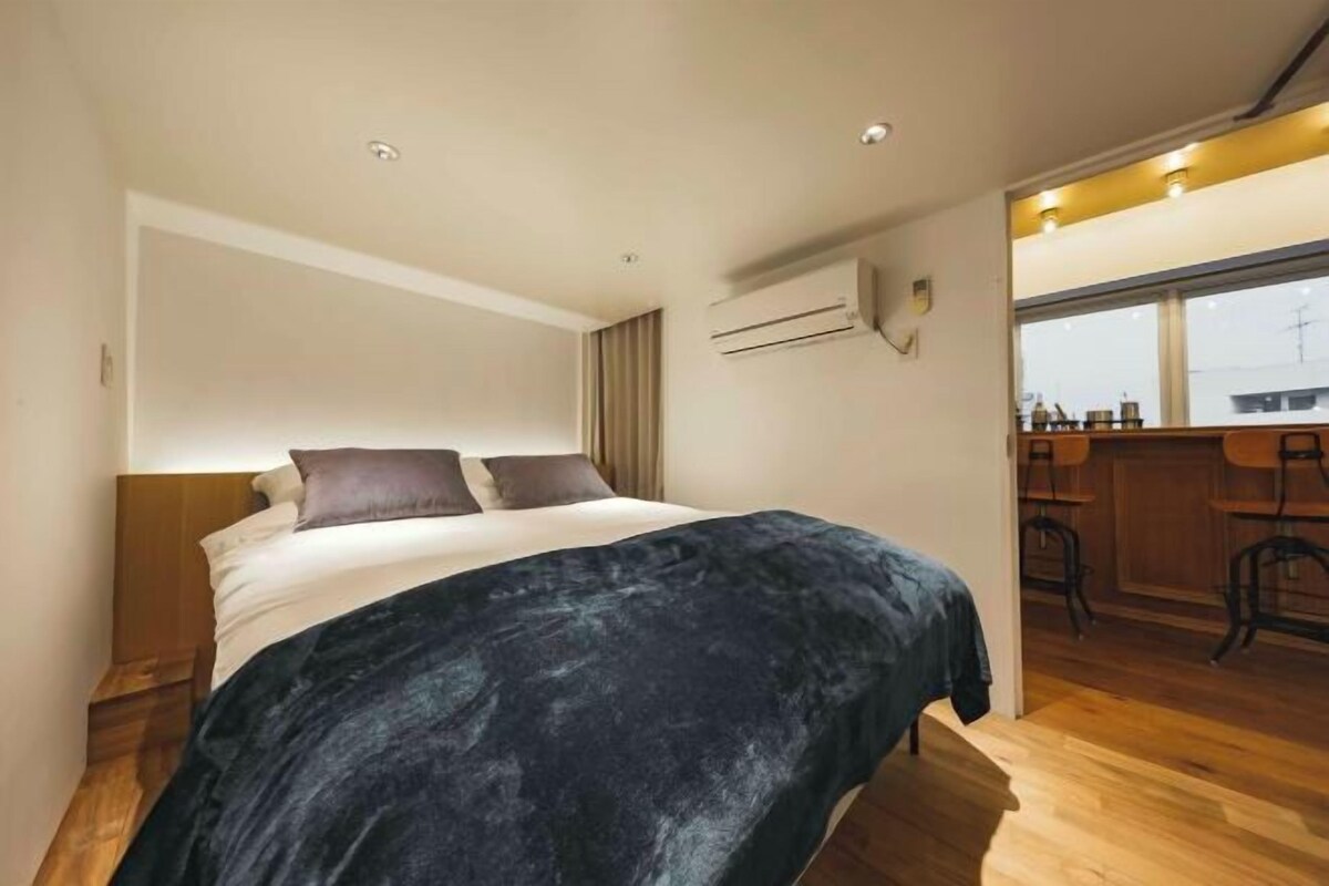 A modern bedroom with a large bed, a wooden headboard, a dark blue blanket, and a window overlooking a cityscape.