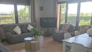 A living room with a sofa, a TV, a dining table, and a view of greenery outside. - Mas Turró - Castelló d'Empúries (Castellón de Ampurias)