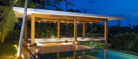 Un área de piscina moderna al aire libre con cubierta de madera, asientos e iluminación ambiental.