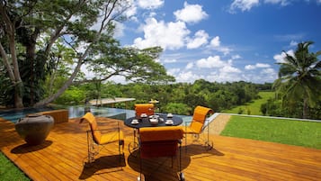Un área de comedor al aire libre con mesa y sillas, con vista a una piscina y abundante vegetación.