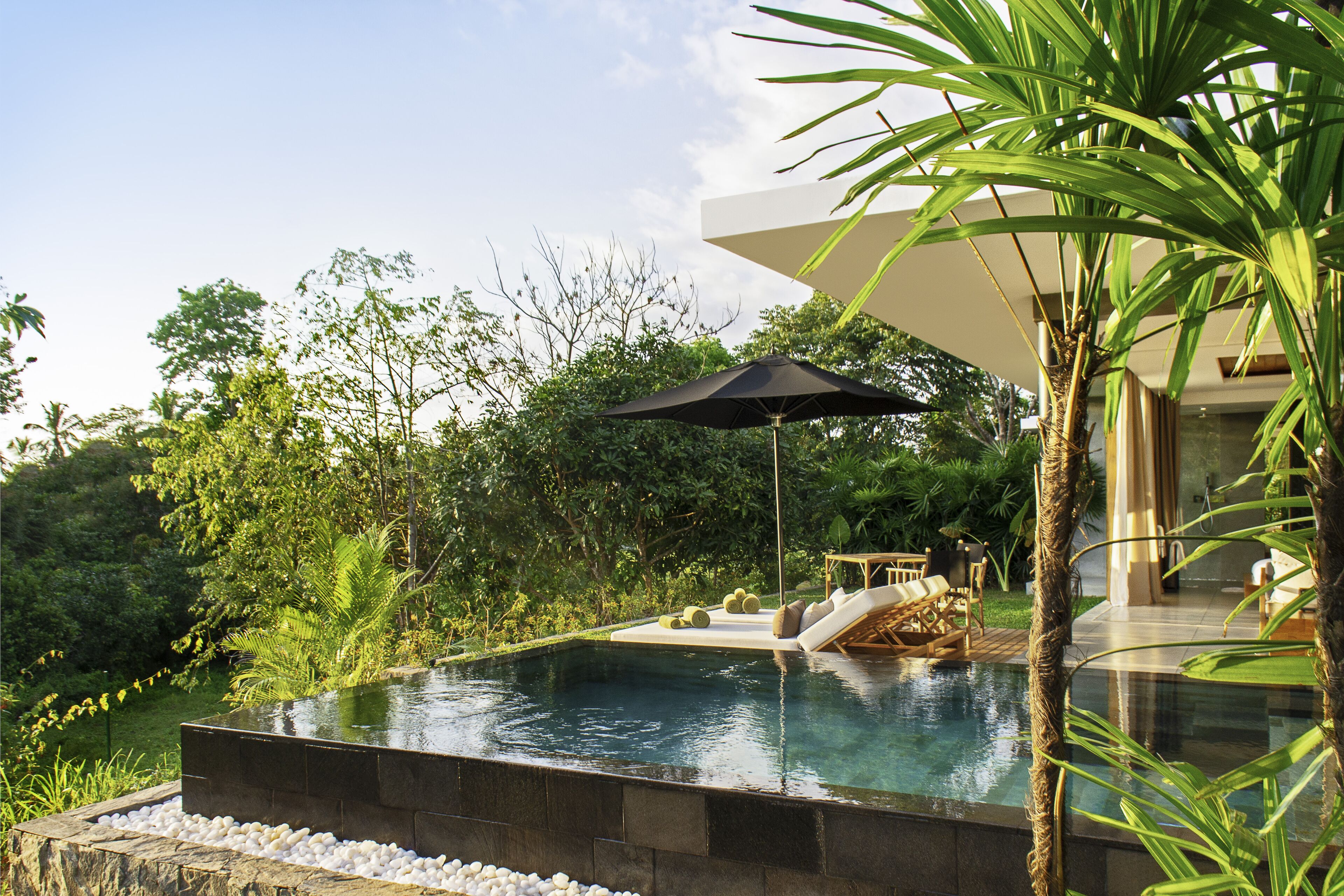 Una piscina con borde de piedra, rodeada de exuberante vegetación y una zona de estar exterior moderna con una sombrilla negra.