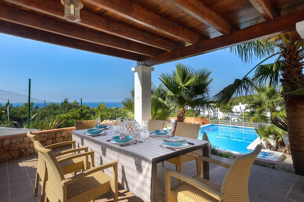 Un área de comedor al aire libre techada con vista a una piscina y al mar.