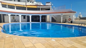 En swimmingpool med fliseterrasse, et overdækket loungeområde og en bygning med altaner i baggrunden.