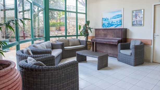 A spacious indoor area with wicker furniture, a piano, and large windows offering views of the outdoors. - HI Pkiin Hostel (Peki'in)