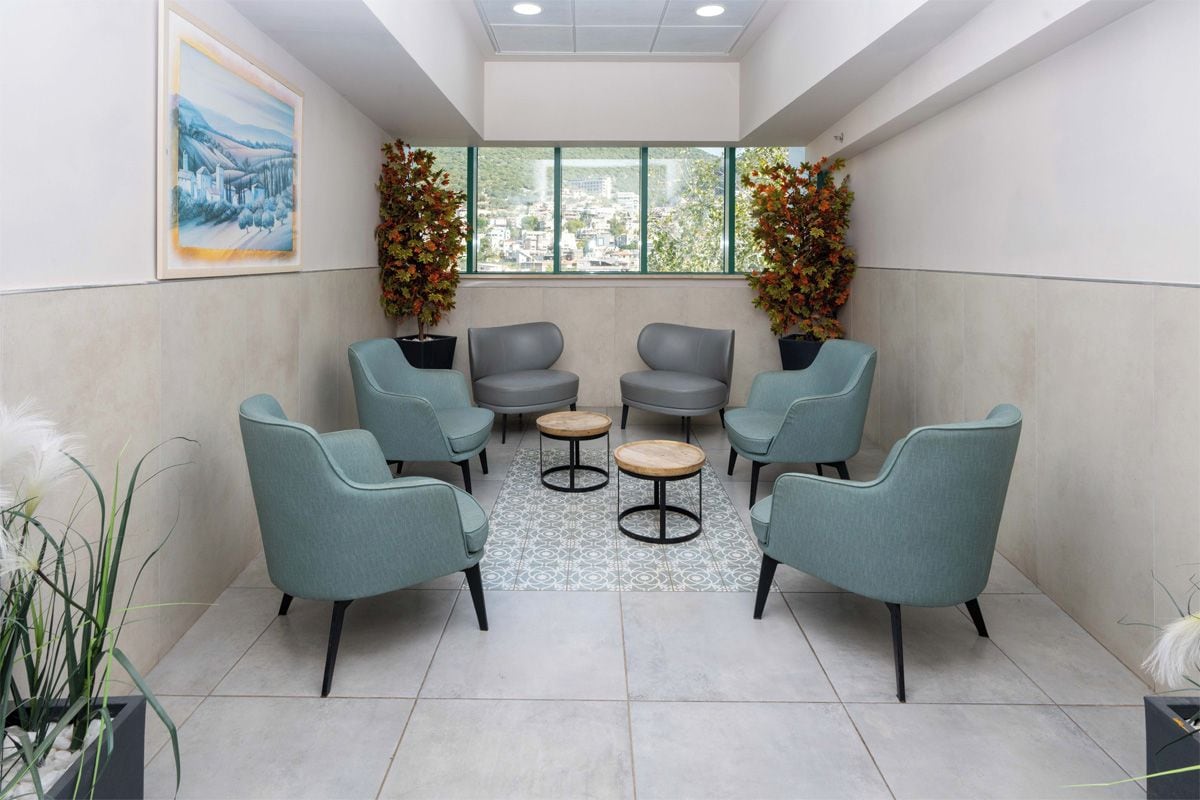 A waiting area with four grey chairs and two small round tables, a large window with a city view, and a framed painting on the wall.