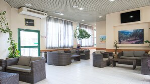 A spacious lobby with wicker furniture, a large window with curtains, a mounted TV, and a painting on the wall. - HI Pkiin Hostel (Peki'in)