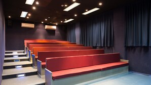 A theater with tiered red seating, a dark stage curtain, and overhead lighting. - HI Pkiin Hostel (Peki'in)