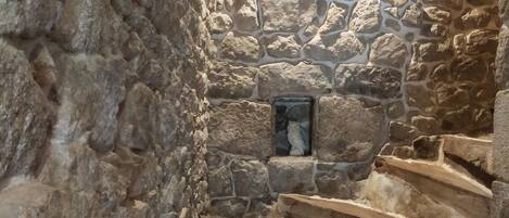 A stone staircase in a narrow, dimly lit corridor with rough stone walls and a small window at the top.