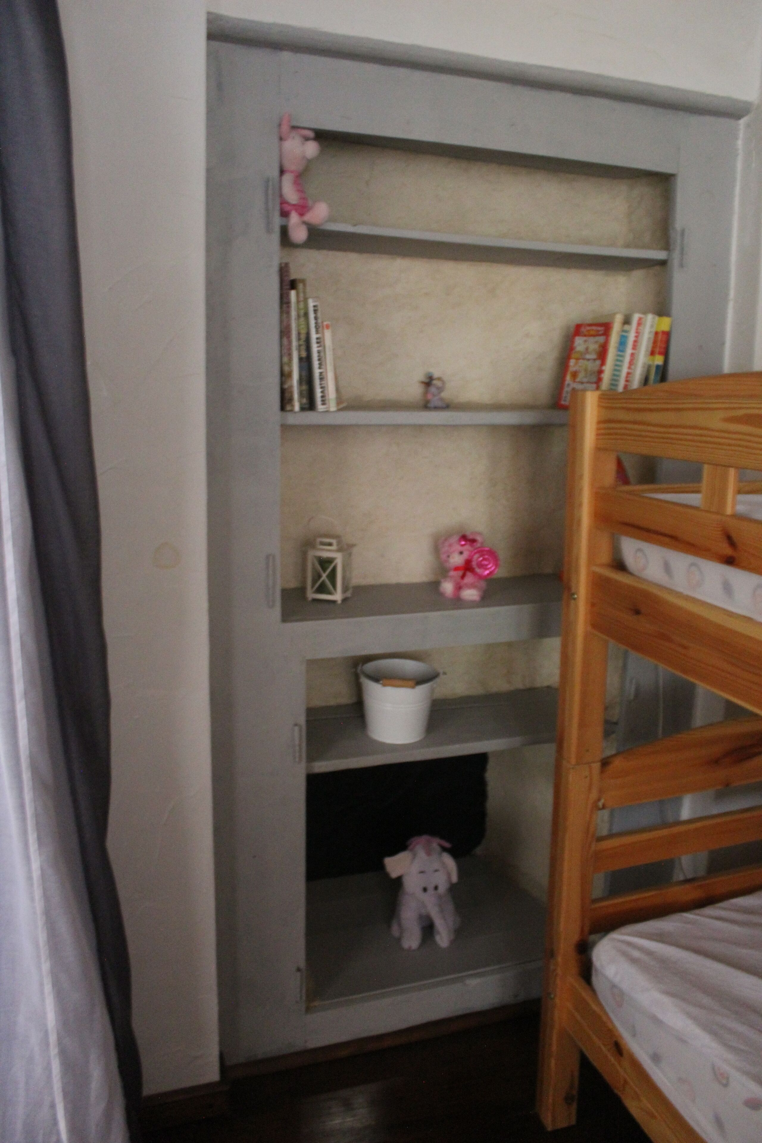 A child's bunk bed with a wooden headboard, a shelf unit with books and toys, and a small white lantern.