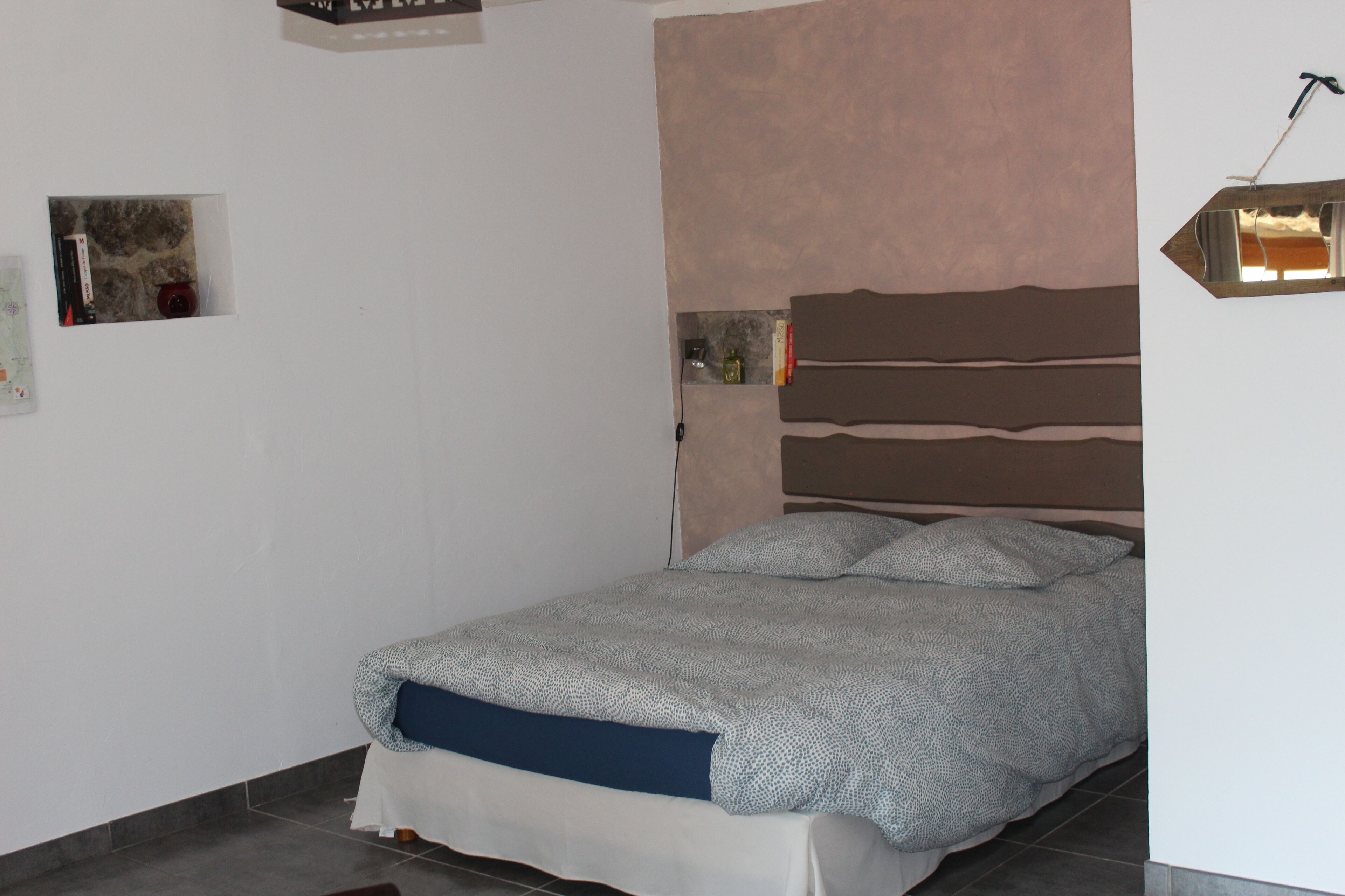 A bedroom with a bed, a headboard, a wall-mounted light fixture, and a small shelf with items.