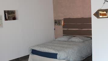 A bedroom with a bed, a headboard, a wall-mounted light fixture, and a small shelf with items.