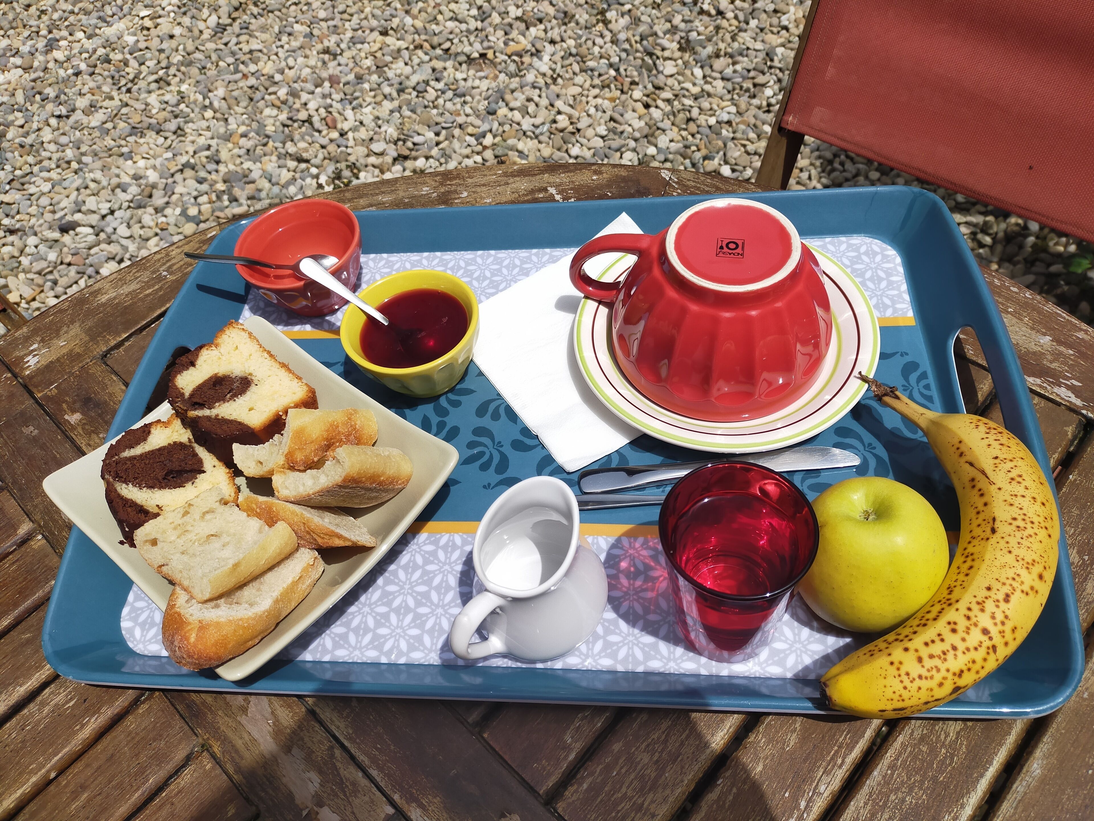 Un plateau avec une théière rouge, une assiette de pain, une banane, une pomme, une tasse de boisson rouge et un petit bol de confiture.