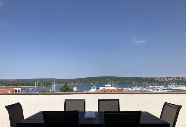 Une table avec quatre chaises et un objet blanc posé dessus, donnant sur une marina avec des bateaux et un paysage vallonné en arrière-plan.