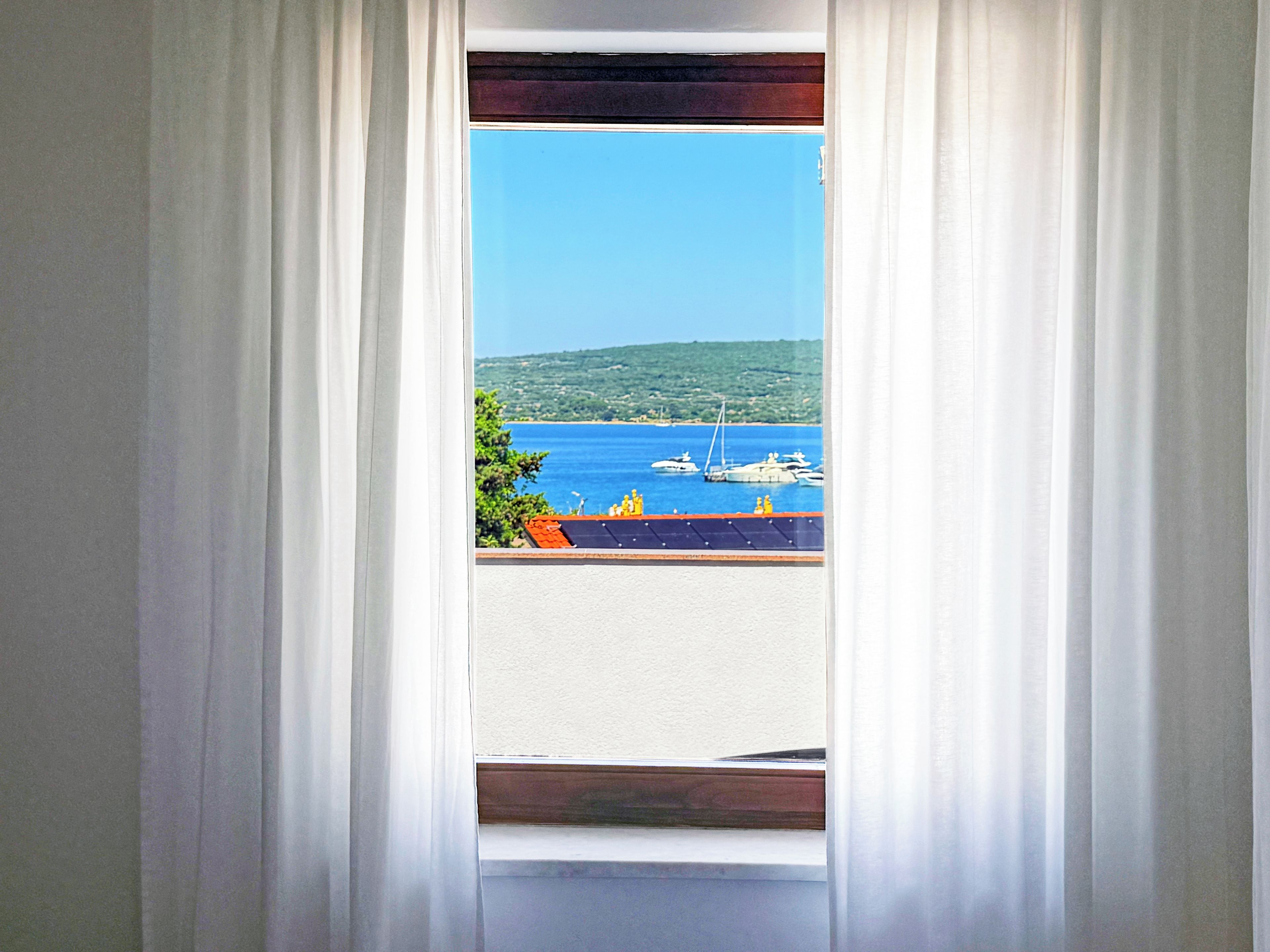 A window with white curtains, a view of a marina with boats, and a hilly landscape in the distance.
