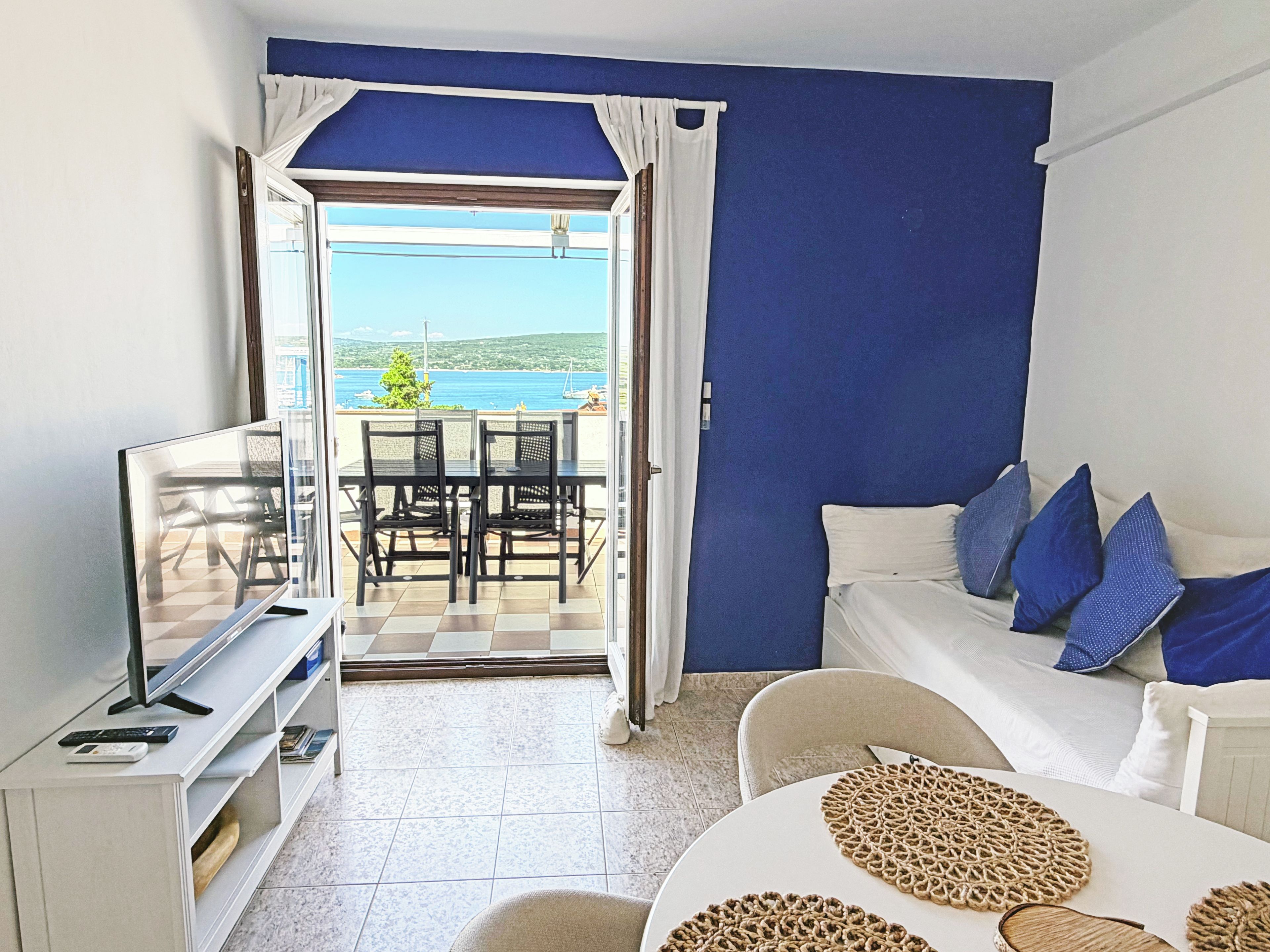 Ein Zimmer mit Fernseher, einem Sofa mit blauen Kissen, einem Essbereich mit Stühlen und Blick aufs Meer.