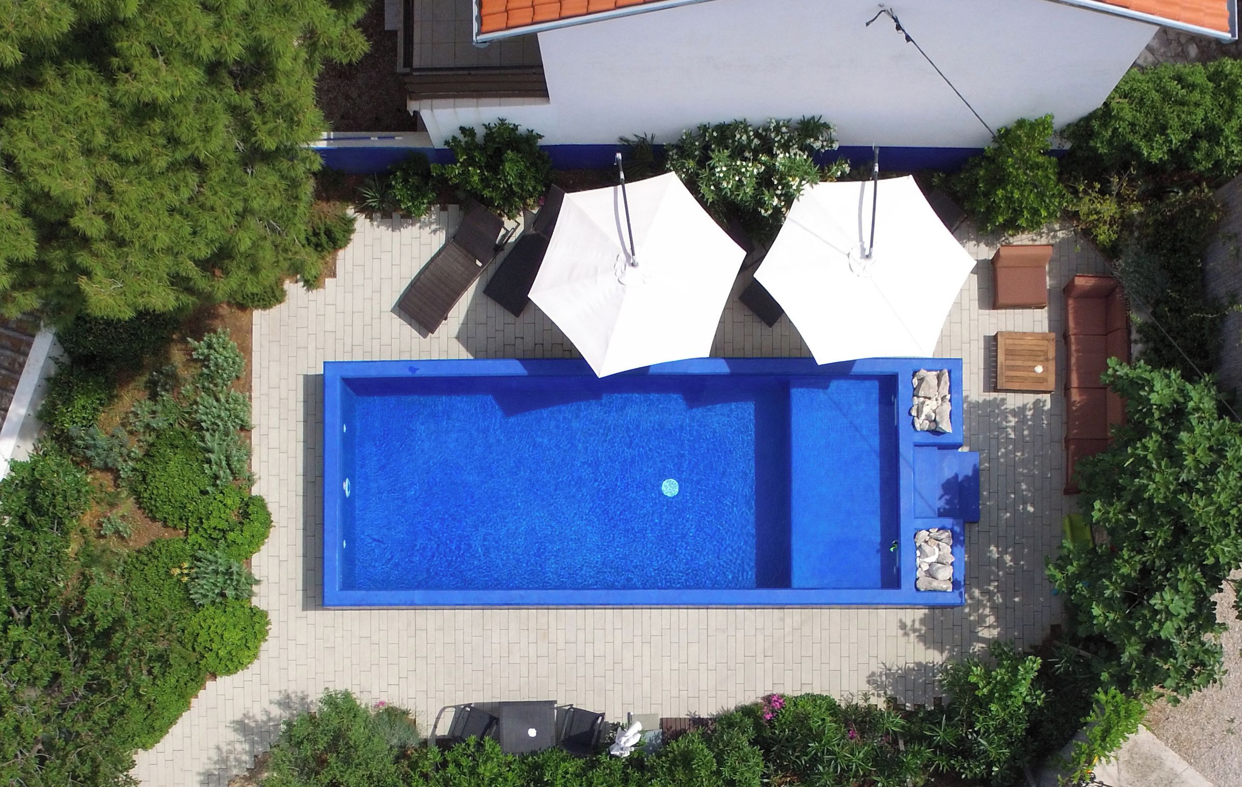 An aerial view of a rectangular swimming pool with two white umbrellas, surrounded by greenery and a paved area.