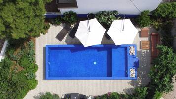 An aerial view of a rectangular swimming pool with two white umbrellas, surrounded by greenery and a paved area.