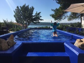 Une personne qui saute dans une piscine, avec la mer et des arbres en arrière-plan.