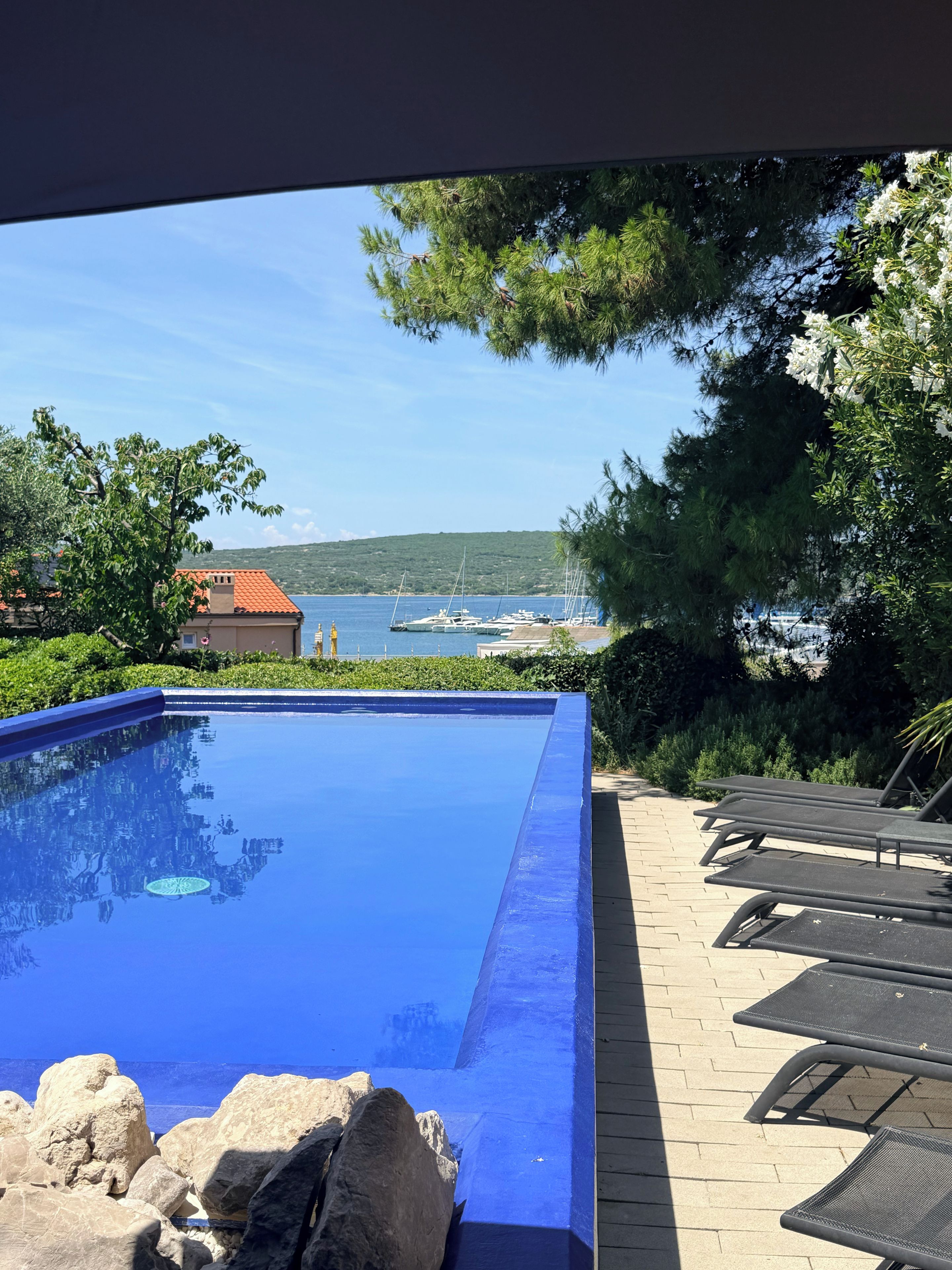 A clear blue swimming pool with lounge chairs and a view of the sea and boats.