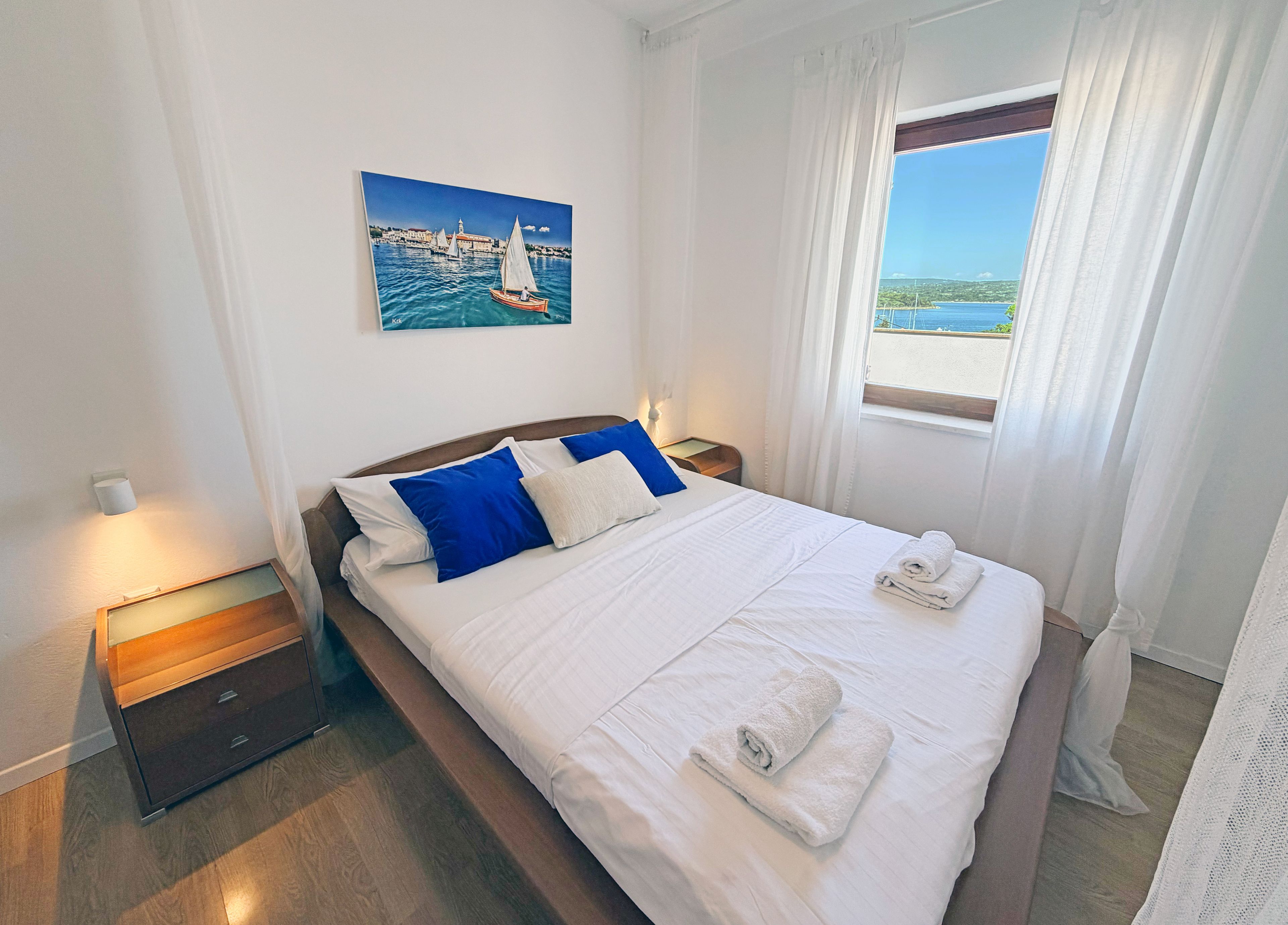 A hotel room with a bed, a nightstand, a bedside table, a window with curtains, and a painting of a coastal scene.