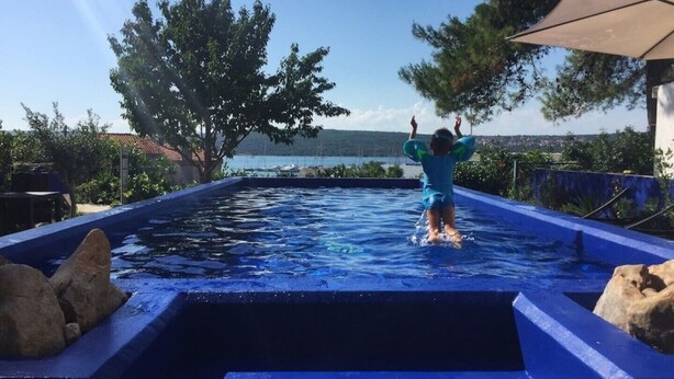 Una persona saltant a una piscina amb vista al mar i als arbres de fons.