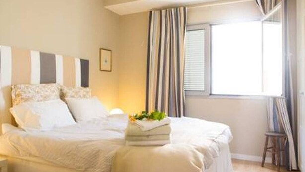 A neatly made bed with a pillow and a folded towel on it, a small stool, and a window with curtains. - Peaceful Top Floor Apt W Balconies-Cambridge RIGHT (AC) (Funchal)