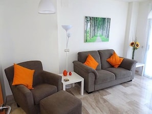 Un salon moderne avec deux canapés gris, chacun agrémenté d’un coussin orange, une table basse blanche et un lampadaire. Un tableau représentant un chemin bordé d’arbres est accroché au mur.