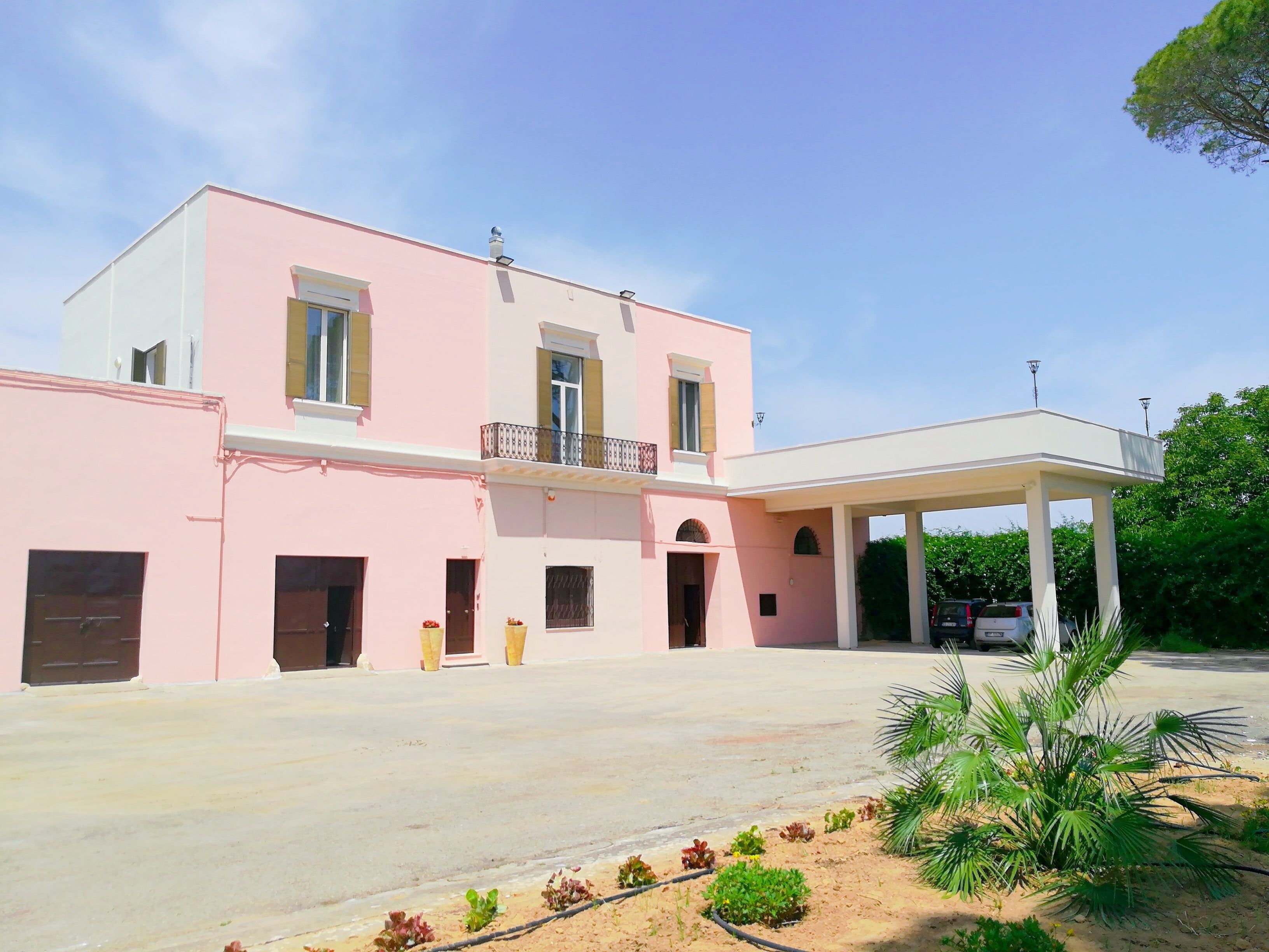 A two-story building with a pink facade, a covered entrance, and a parking area with a few cars.