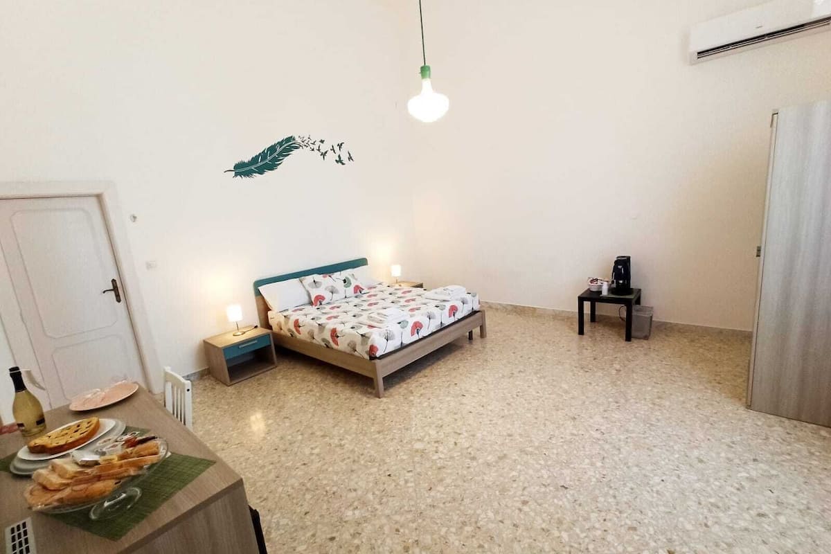 A bedroom with a bed, bedside tables, a desk with food, and an air conditioning unit.