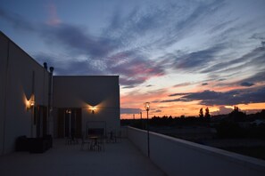 A rooftop terrace with outdoor seating, a building with a flat facade, and a sunset sky. - Calle Dei Saraceni (Carmiano)
