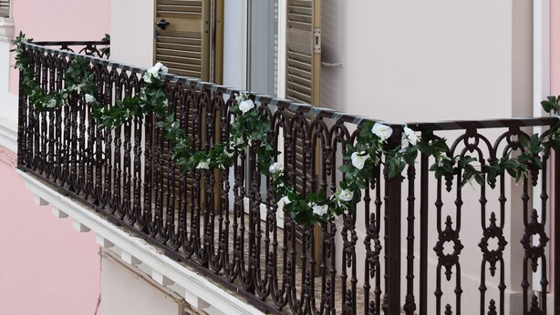 Un balcón con barandilla de hierro forjado, una ventana con contraventanas y una pared rosa.