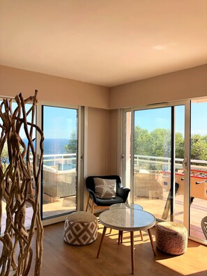 A modern living room with a glass door leading to a balcony, a black armchair, a round glass table, and two patterned ottomans. - Large family villa with an exceptional view of the Calanques—sleeps 10 (Ensuès-la-Redonne)