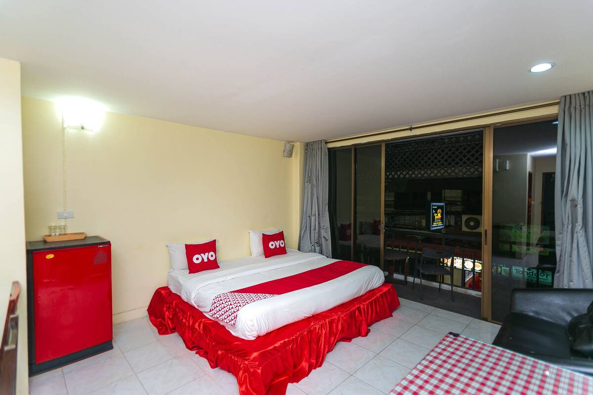 A hotel room with a bed, a red minibar, a table with a checkered tablecloth, and a sliding glass door leading to a balcony.